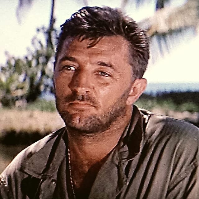 Robert Mitchum in close-up against a tropical backdrop of palms and sea